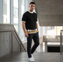 Load image into Gallery viewer, A man wearing a black short-sleeve ringer t-shirt with white trim, black pants, and white sneakers, walking in a modern concrete hallway.